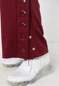 Bordowe spodnie do kostek z dużymi, okrągłymi guzikami po bokach, skomponowane z białymi butami sportowymi z przezroczystą podeszwą.