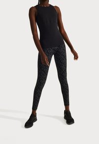 Černý bezrukávový sportovní top spárovaný s těsnými legínami s tmavým geometrickým vzorem. Modelka stojí s jednou nohou vpřed a má na sobě černé tenisky.