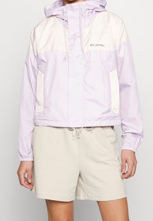 Personne portant une veste à capuche Columbia violet clair et crème et un short beige, debout avec les bras détendus le long du corps.