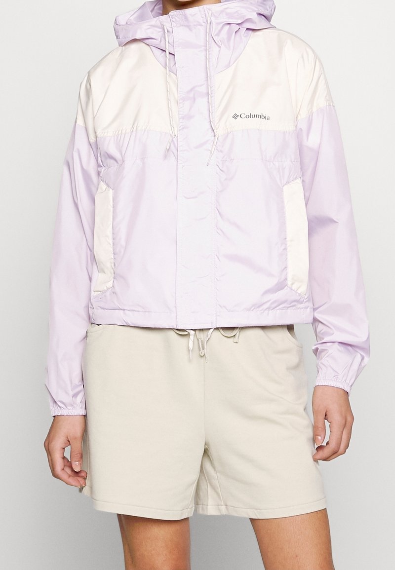 Personne portant une veste à capuche Columbia violet clair et crème et un short beige, debout avec les bras détendus le long du corps.