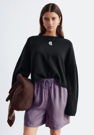Kvinde iført sort oversized sweater, blå og brune stribede shorts, holder brun strikket taske med tørklæde bundet til, neutralt baggrund.