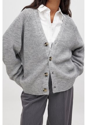 Femme portant un cardigan en maille gris épais avec de gros boutons, par-dessus une chemise blanche à col, et un pantalon gris ample.