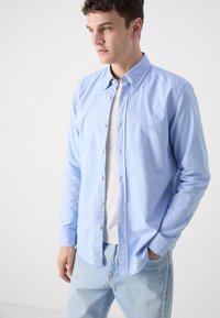 Lichtblauw button-up shirt van katoen met lange mouwen, een button-down kraag en geborduurd logo op de borst. Draag het over een wit t-shirt.