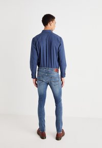 Camisa de mezclilla abotonada en azul oscuro, con un corte ajustado y manga larga. Combinada con unos jeans slim-fit en azul claro y zapatos marrones, mostrando un detalle mínimo.