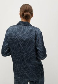 Donna con capelli castano chiaro raccolti in uno chignon, che indossa una camicia a maniche lunghe blu scuro con motivo e pantaloni abbinati, rivolta di spalle su uno sfondo semplice.