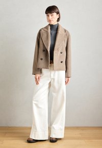 Veste double boutonnage en mélange de laine marron avec de grands revers, portée sur un col roulé gris, associée à un pantalon large blanc et des chaussures marron.