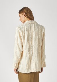 Blazer color crema con rayas verticales texturizadas. Presenta un cuello con muesca, mangas largas y bordes deshilachados. Detalle de botones en los puños.