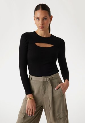 Black ribbed long-sleeve top with a cutout design at the neckline, paired with light olive green trousers featuring a tied waist.