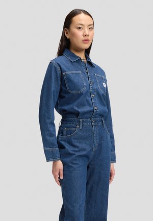 Ung kvinde iført en blå denimjakke og matchende højtaljede jeans står mod en ensartet lys baggrund.