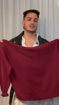 Άνδρας με κοντά σκούρα μαλλιά και γενειάδα κρατά ένα μωβ πουλόβερ μπροστά σε λευκό φόντο κουρτίνας.