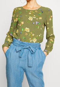 Zielona kwiatowa bluzka z długimi rękawami, okrągłym dekoltem i wyrazistymi wzorami kwiatowymi; zestawiona z jasnymi dżinsowymi spodniami o wysokim stanie i muszką.