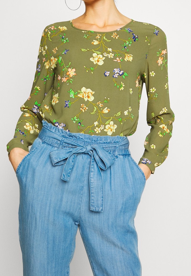 Zielona kwiatowa bluzka z długimi rękawami, okrągłym dekoltem i wyrazistymi wzorami kwiatowymi; zestawiona z jasnymi dżinsowymi spodniami o wysokim stanie i muszką.