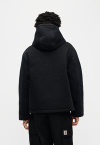 Μαύρο μπουφάν με κουκούλα από υφασμάτινη υφή, με άνετη εφαρμογή, ορατές ραφές και ελαφριά γυαλάδα. Το μπουφάν εμφανίζεται από την πίσω πλευρά.