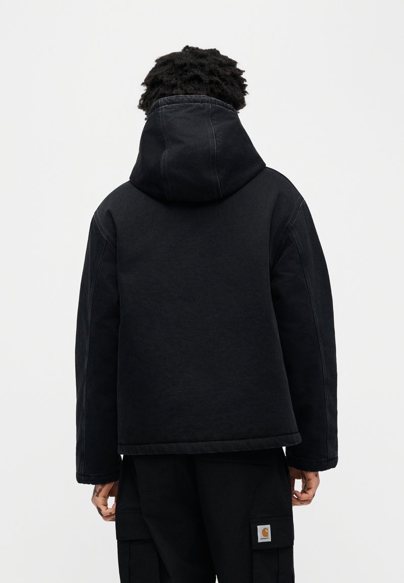 Veste noire à capuche en tissu texturé, avec une coupe décontractée, des coutures visibles et un léger éclat. La veste est présentée de dos.