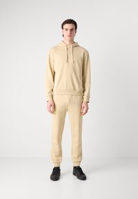 Hanorac bej și pantaloni sport asortați, confecționați dintr-o țesătură moale. Dispune de un căpăcel cu șnur, manșete ribbrate și teniși negri. Design minimalist.
