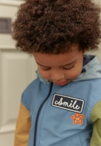 Jas voor kinderen in blauwe, groene en gele panelen; met een "glimlach" patch en stervormige accenten. Het materiaal lijkt lichtgewicht en gestructureerd.
