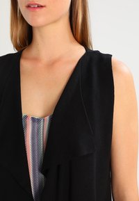 Black sleeveless vest with a draped front, made of soft fabric. Underneath, a multicolored patterned top with stripes in pink, blue, and white.