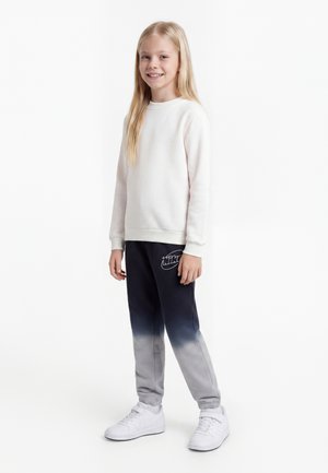Smilende pige med langt blondt hår iført hvid sweatshirt, sorte til grå gradient sweatpants med logo og hvide sneakers, stående mod hvid baggrund.