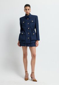 Granatowa sukienka typu żakietowego o fakturowanej tkaninie, z wysokim kołnierzem, złotymi guzikami, dwoma przednimi kieszeniami oraz regulowanym paskiem na talii. Połączona z brązowymi szpilkami.