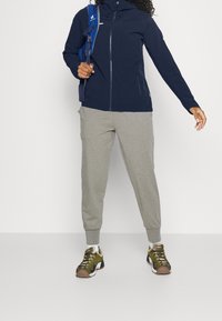 Navy softshell jacket with a front zipper, paired with light gray sweatpants and multicolored sneakers. Jacket features a hood and pockets.