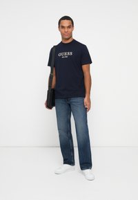 Man staat met een marineblauwe Guess t-shirt, blauwe jeans, witte sneakers en draagt een zwarte schoudertas tegen een effen witte achtergrond.