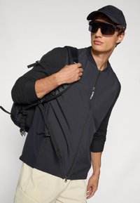 Black zip-up jacket with contrasting sleeves, worn over a light-colored outfit; complemented by a black cap and oversized sunglasses.