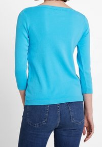 En person står med ryggen synlig, klädd i en ljusblå långärmad tröja och mörkblå jeans, framför en enkel vit bakgrund.