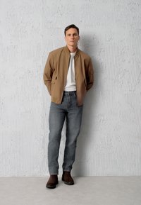 Brązowa kurtka bomberka na białej koszulce, w połączeniu z szarymi dżinsami i brązowymi zamszowymi butami. Proste tło z teksturowaną jasnoszaro ścianą.