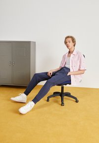 Rozā svītrainā krekls ar zaļiem akcentiem, pelēkie treniņbikses un baltas tenisa kedas uz melna ergonomiska krēsla pretī dzeltenai grīdai.