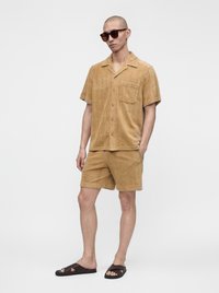 Homme portant une chemise beige à manches courtes boutonnée avec un short assorti, des lunettes de soleil foncées et des sandales slide foncées, debout avec une main dans la poche.