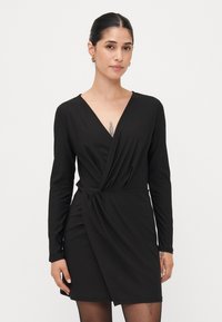 Abito nero avvolgente realizzato in un tessuto morbido e elastico. Presenta maniche lunghe, una profonda scollatura a V e dettagli arricciati in vita.