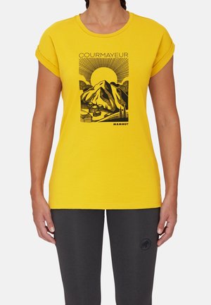Camiseta amarilla con mangas remangadas que muestra un gráfico negro de montañas, sol y el texto "COURMAYEUR", combinada con pantalones oscuros.