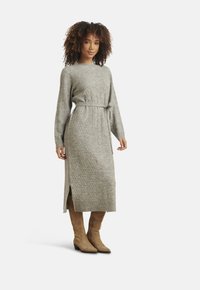 Donna con capelli ricci che indossa un vestito midi grigio in maglia con cintura e stivali in camoscio marrone chiaro sopra il ginocchio, in piedi su uno sfondo bianco.