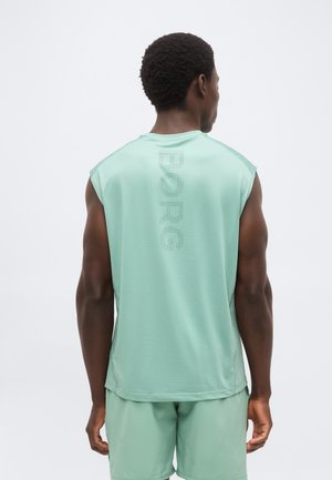 Muž v nosení mintovo zeleného športového tielka bez rukávov s perforovaným textom "BORG" na chrbte a zladenými šortkami, stojí chrbtom ku kamere.