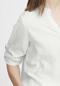 Blouse en lin blanc avec une texture douce, dotée d'un décolleté en V et de manches retroussées. Détails de couture distinctifs au niveau des poignets.