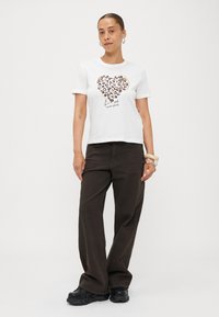 Weißes Baumwoll-T-Shirt mit einem Herzdesign aus Leopardenmuster und schwarzem Text "Liebe scheitert nie." Kombiniert mit dunklen, weit geschnittenen Hosen.