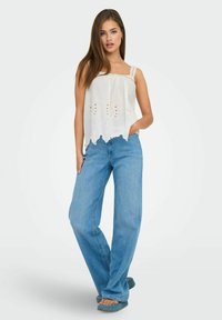 Blusa bianca ricamata con orlo a scallop e spalline sottili, abbinata a jeans azzurri a gamba larga e sandali blu verde a slip-on.