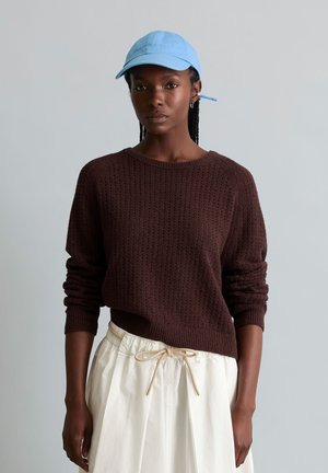 Vrouw met een blauwe pet, donkerbruine textuurtrui en een off-white rok met trekkoord, staand voor een effen lichtgrijze achtergrond.