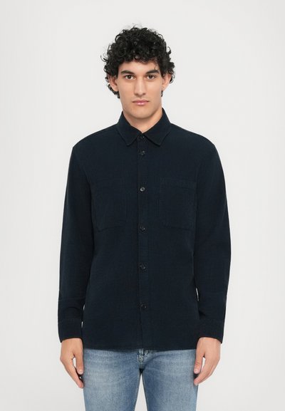 Chemise bleue marine à boutons en tissu texturé, avec deux poches avant, un col classique et des manches longues, associée à un jean bleu clair.