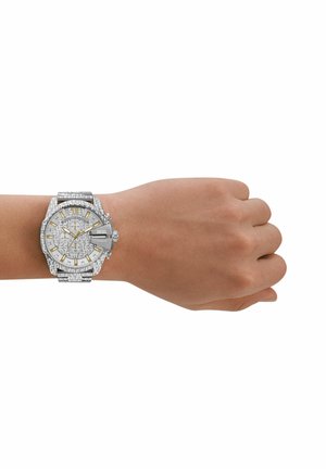 Handgelenk mit silberner Diesel Chronograph-Uhr mit gemustertem Armband, goldenen Akzenten und mehreren Zifferblättern an einer hellen Haut.