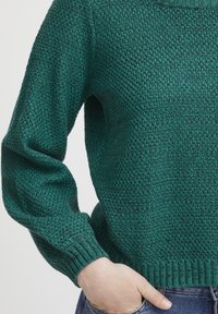 Grüner Strickpulli mit texturiertem Muster, gerippten Bündchen und Rundhalsausschnitt; präsentiert mit Händen in den Taschen und Jeans.