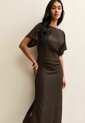 Femme aux longs cheveux foncés portant une robe marron foncé cintrée avec des manches courtes flottantes et des bracelets dorés, debout contre un mur beige.