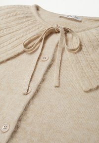 Cardigan beige en tricot avec motif torsadé sur les épaules, fermeture à boutons et nœud noué au col rond.