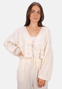 Cremefarbenes, kurzes Oberteil mit gewelltem Spitzenbesatz und Bindeschluss; lange, schwingende Ärmel; über einem hellen Trägershirt getragen.