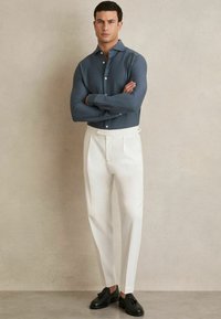 Camisa gris de botones con textura y pantalones blancos a medida, acompañados de zapatos negros. El modelo está de pie con los brazos cruzados frente a un fondo neutro.