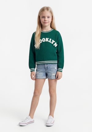 Jeune fille aux longs cheveux blonds portant un pull vert « Brooklyn », un short en denim et des baskets blanches, debout devant un fond blanc uni.