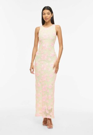 Femme portant une robe longue beige sans manches avec des appliqués en dentelle florale rose, debout et face à l'avant sur un fond blanc uni.