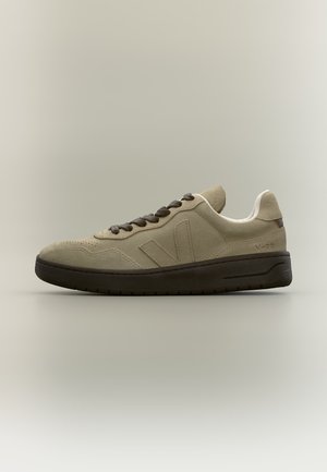 Baskets en daim beige avec semelle et lacets marron foncé, logo latéral et col rembourré sur fond neutre.