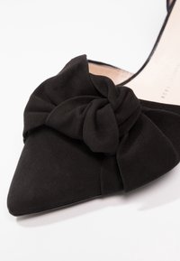 Chaussure en daim noir à bout pointu avec un grand nœud en tissu torsadé sur l'avant et un design latéral ouvert.