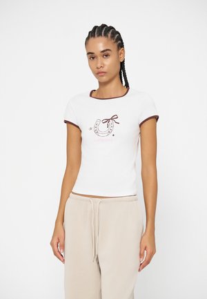 Jeune femme avec des tresses portant un t-shirt court blanc avec un motif fer à cheval et un pantalon beige à cordon, debout devant un fond blanc.